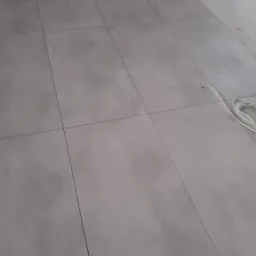 Ułożone szare płytki podłogowe z ciemnymi fugami, widoczny fragment kabla oraz młotek i płytka w rogu pomieszczenia.