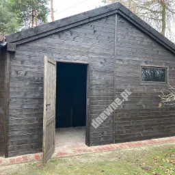 Ciemna, drewniana altana z otwartymi drzwiami, widoczna ceglana podmurówka i fragment dachu sąsiedniego budynku.