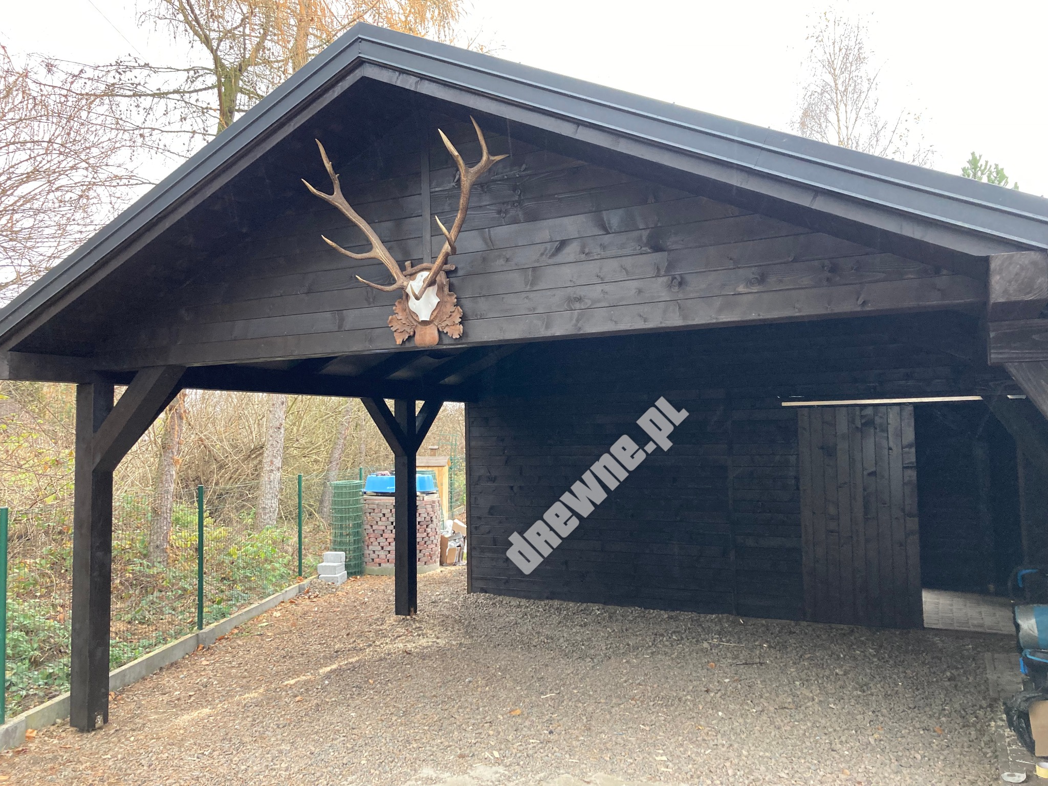 Ciemna, drewniana wiata garażowa z dekoracją z poroża jelenia na ścianie frontowej, widok z zewnątrz na żwirowe podłoże i ogrodzenie z siatki.