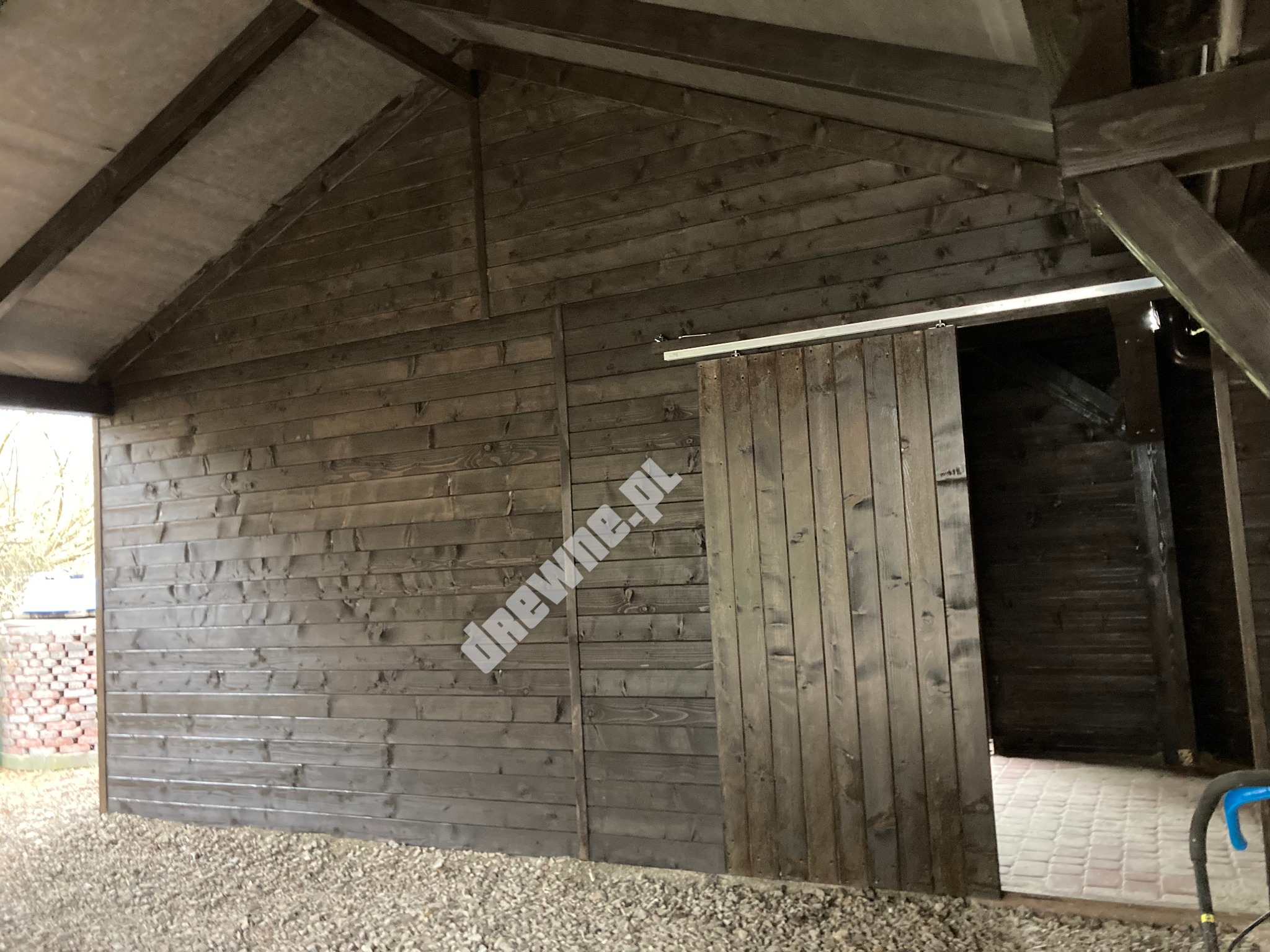 Zadaszona konstrukcja z ciemnego drewna z przesuwnymi drzwiami, widok na wnętrze z wyłożoną kostką podłogą, otoczenie z kamienistym podłożem.