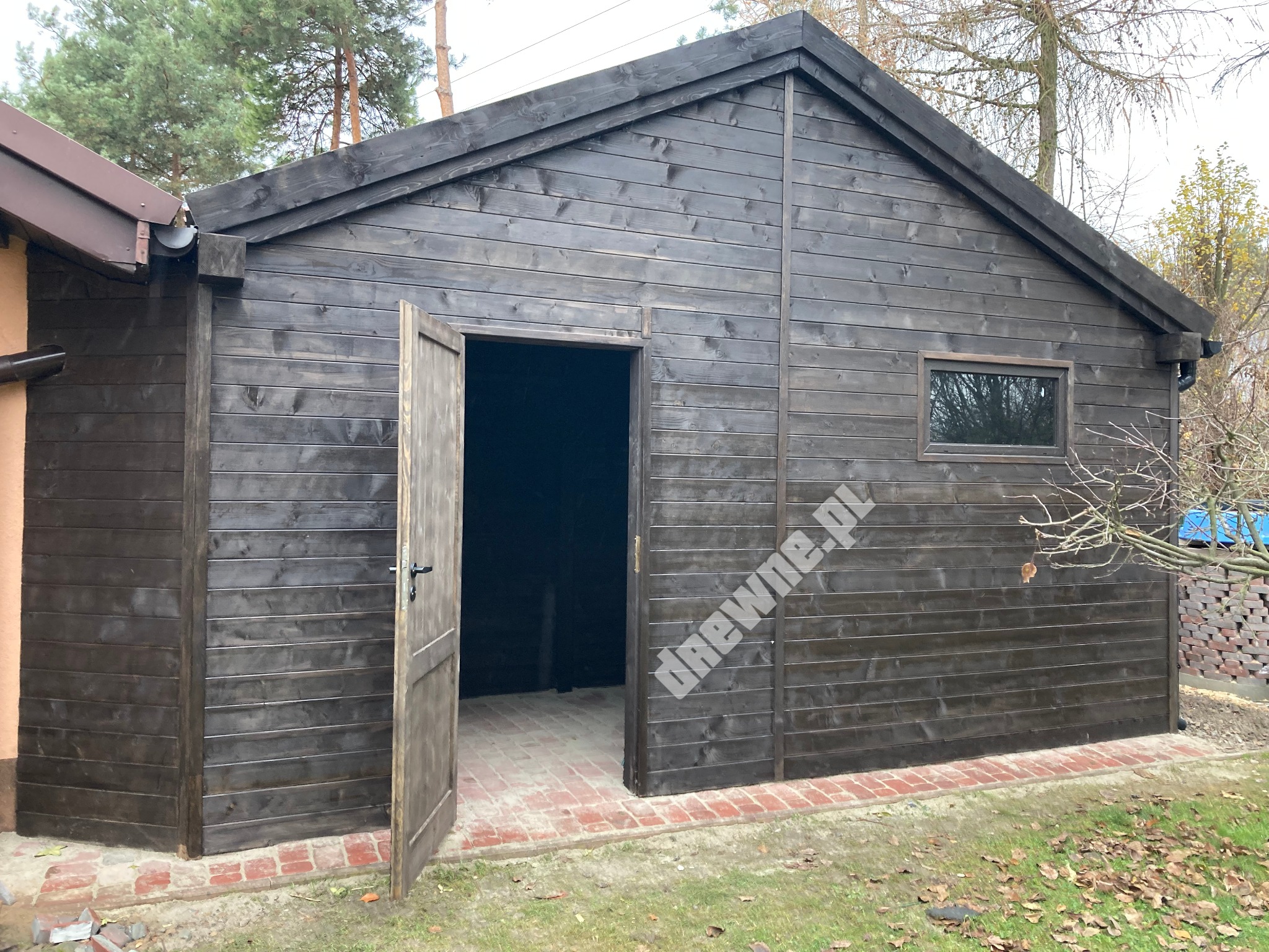 Ciemna, drewniana altana z otwartymi drzwiami, widoczna ceglana podmurówka i fragment dachu sąsiedniego budynku.