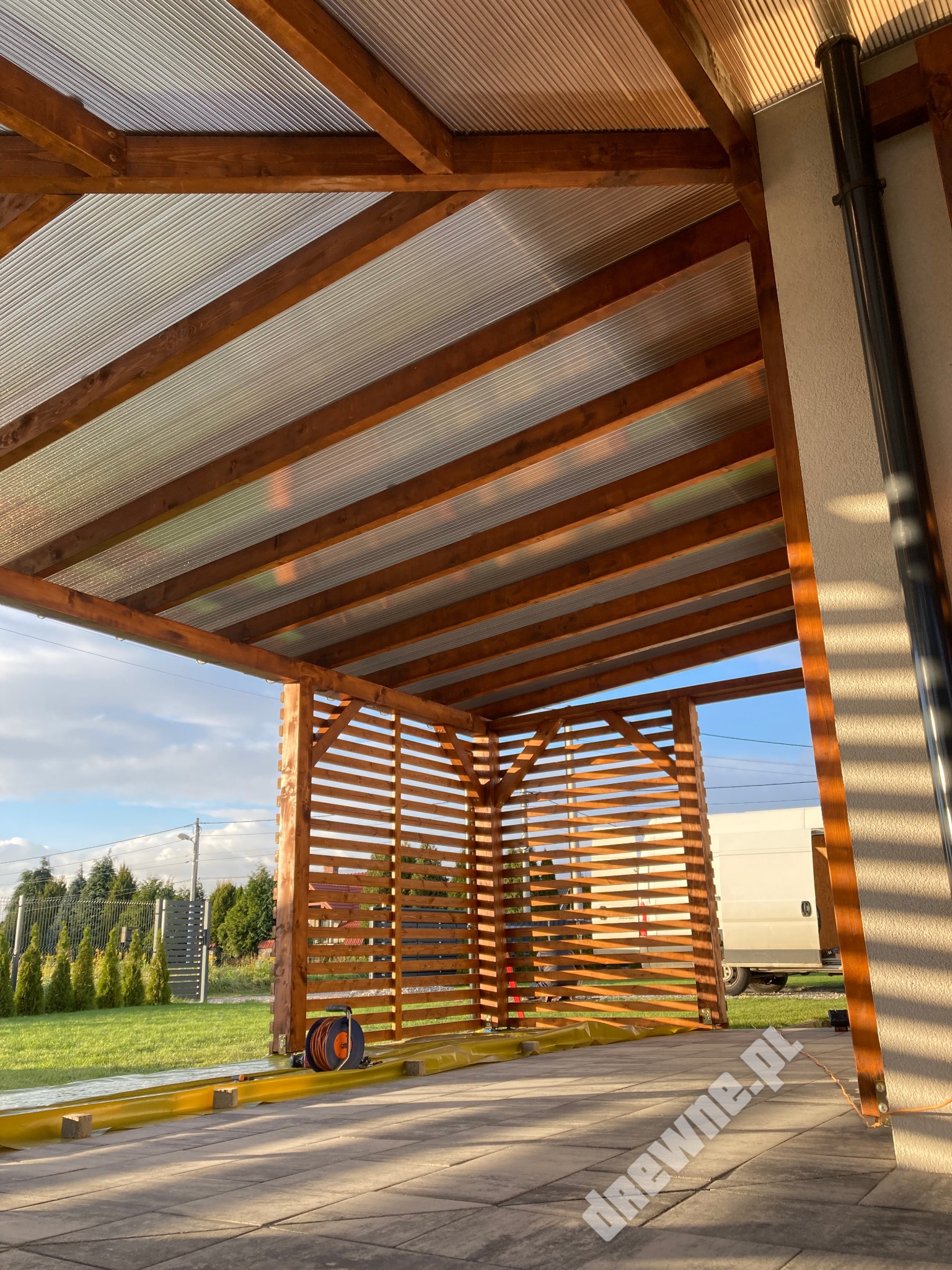 Drewniana konstrukcja altany z transparentnym zadaszeniem i ażurowymi ścianami w trakcie montażu, widoczna rozłożona folia ochronna na podłożu.