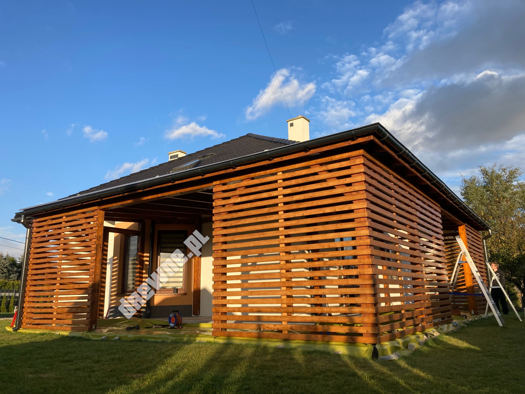 Nowoczesna, drewniana konstrukcja osłaniająca taras domu jednorodzinnego z widoczną drabiną i elementami zabezpieczającymi podczas montażu, na tle błękitnego nieba z chmurami.