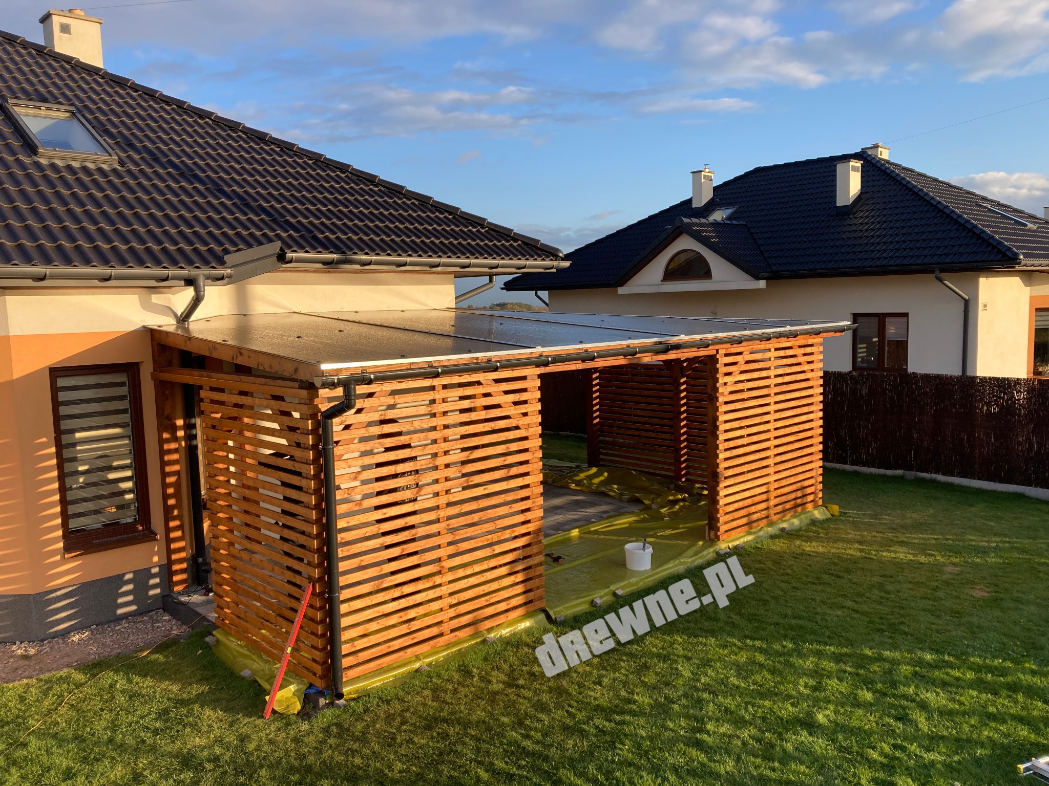 Nowoczesna altana z drewnianymi lamelami, przylegająca do domu z widocznym poziomem, zabezpieczona folią budowlaną, w tle domy z ciemnymi dachami