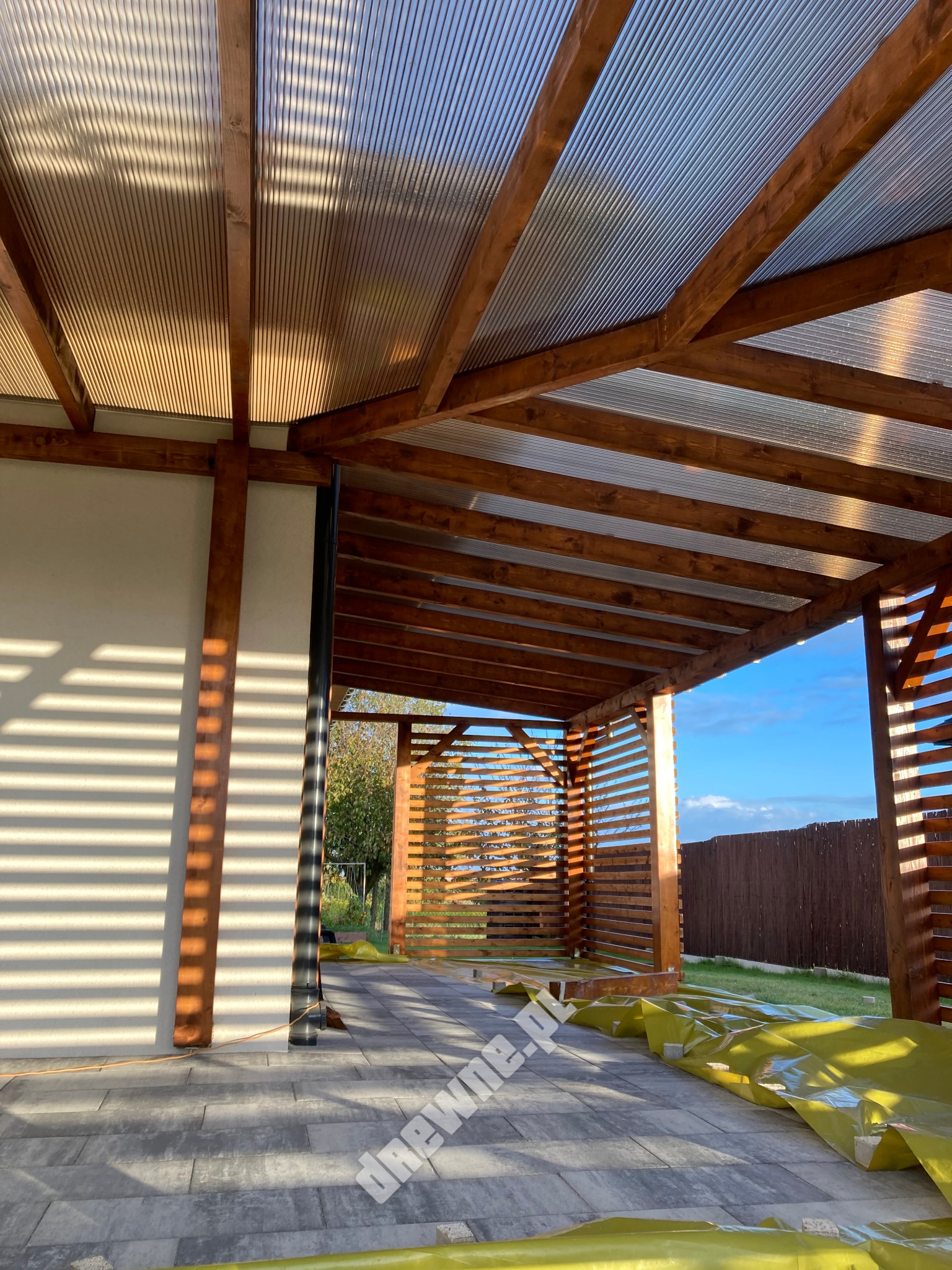 Nowoczesna altana z drewnianą konstrukcją i przezroczystym zadaszeniem, w trakcie budowy, zabezpieczona folią ochronną na podłożu z kostki brukowej, oświetlona popołudniowym słońcem rzucającym...