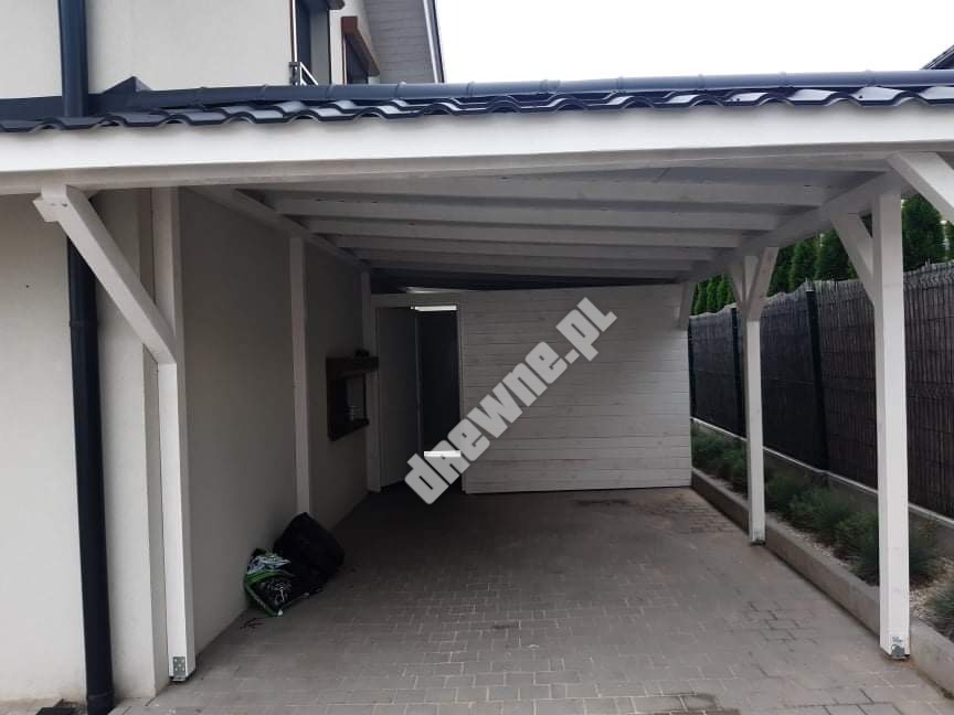 Biała, drewniana wiata garażowa przylegająca do domu, z czarnym dachem i podjazdem z kostki brukowej. Widoczne elementy konstrukcyjne i wykończeniowe.