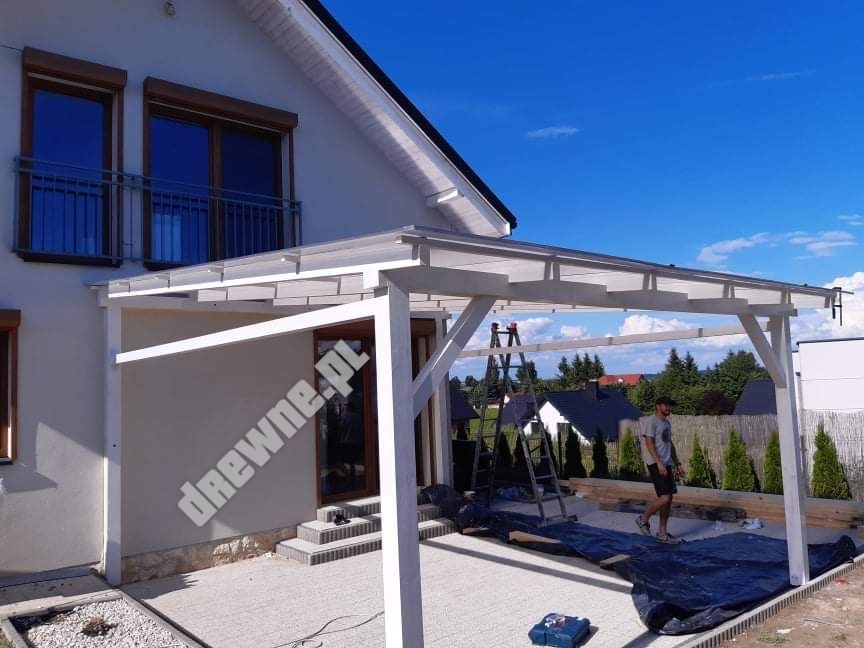 Biała, drewniana konstrukcja z przezroczystym zadaszeniem w trakcie montażu na betonowym patio, widoczna drabina i pracownik w tle, błękitne niebo.