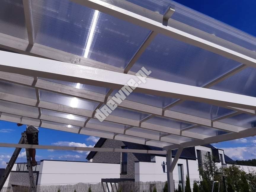 Biała konstrukcja z przezroczystym zadaszeniem z poliwęglanu, widoczna z dołu, z pracownikiem na drabinie i domem w tle.