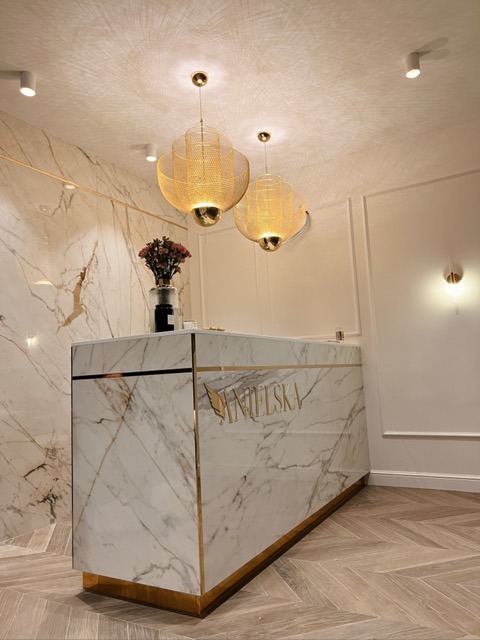 Recepcja z marmurowym blatem i złotymi akcentami, oświetlona dwoma ażurowymi lampami wiszącymi w nowoczesnym wnętrzu z parkietem w jodełkę.