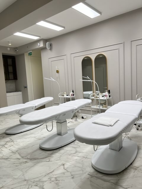 Jasne wnętrze gabinetu kosmetycznego z trzema białymi leżankami na marmurowej podłodze, oświetlone lampami LED i naturalnym światłem wpadającym przez okno.