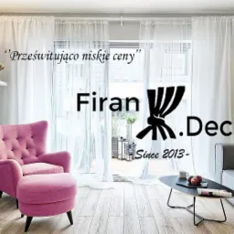Jasne, zwiewne zasłony z logo firmy 'Firan.Decor' na tle balkonu, w salonie z różowym fotelem i szarą sofą.