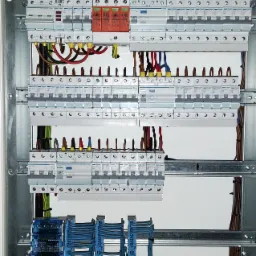 Otwarta skrzynka elektryczna z zamontowanymi zabezpieczeniami Hager, widoczne kable w różnych kolorach uporządkowane wewnątrz, listwy montażowe i bezpieczniki różnego typu.