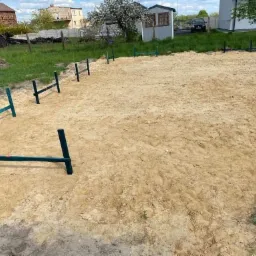 Przygotowany teren pod budowę z wyznaczonym obrysem fundamentów za pomocą zielonych palików i poprzeczek, widok z góry, w tle domy jednorodzinne i zielona trawa.