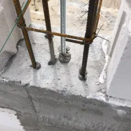 Fragment ściany z betonu komórkowego z widocznym zbrojeniem pionowym i poziomym, oraz gwintowanym prętem stalowym. Zbrojenie związane drutem. Widoczne niedociągnięcia w wypełnieniu spoin między...