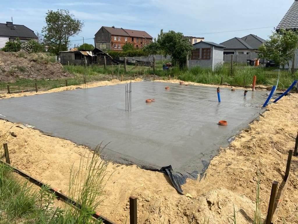 Wylany fundament pod budowę domu jednorodzinnego, z widocznymi pionowymi prętami zbrojeniowymi i rurami kanalizacyjnymi wystającymi z betonu, otoczony piaskiem i drewnianymi szalunkami.