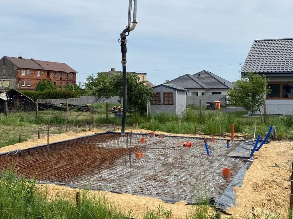 Wylewanie betonu na zbrojenie fundamentu pod budowę domu jednorodzinnego, widoczne rury kanalizacyjne i pompa do betonu na tle zabudowy mieszkalnej.