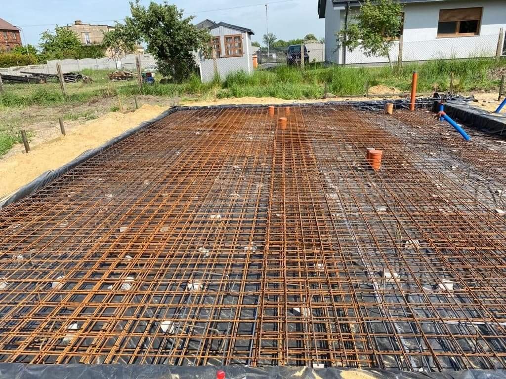 Zbrojenie fundamentów domu jednorodzinnego, widoczne stalowe pręty tworzące siatkę na czarnej folii izolacyjnej, z pomarańczowymi rurami kanalizacyjnymi, teren budowy w słoneczny dzień.