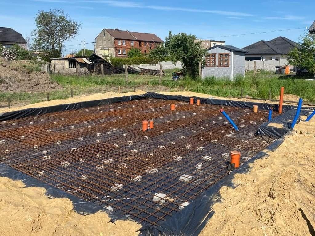 Zbrojenie fundamentu w kwadratowym wykopie, z widocznymi rurami kanalizacyjnymi i folią izolacyjną, na tle zabudowy jednorodzinnej w słoneczny dzień.