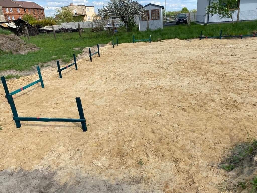 Przygotowany teren pod budowę z wyznaczonym obrysem fundamentów za pomocą zielonych palików i poprzeczek, widok z góry, w tle domy jednorodzinne i zielona trawa.