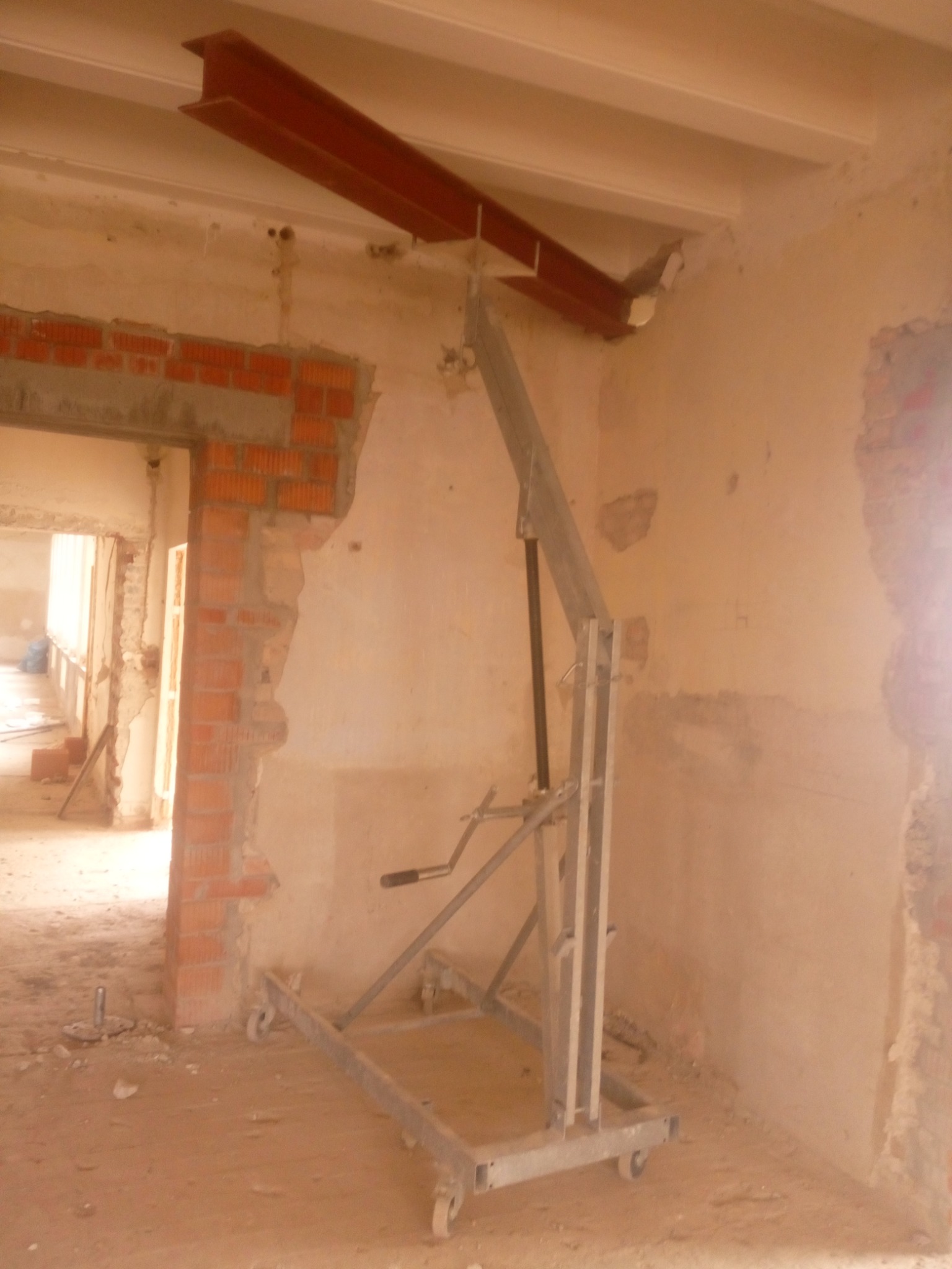 Wnętrze w trakcie prac budowlanych z widoczną stalową belką podtrzymującą strop, podnośnikiem na kółkach i fragmentem ściany z czerwonej cegły przy otworze drzwiowym.