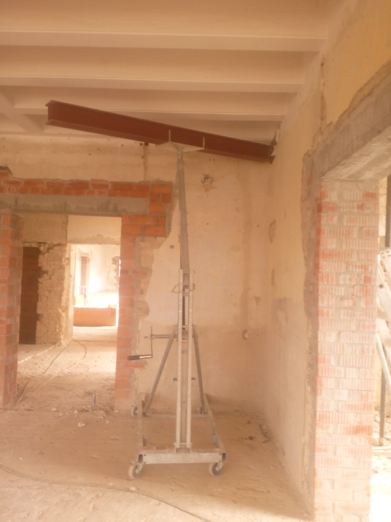 Wnętrze budynku w trakcie prac konstrukcyjnych, widoczny stalowy profil podtrzymywany przez podnośnik na tle surowych ścian z cegły i betonu.