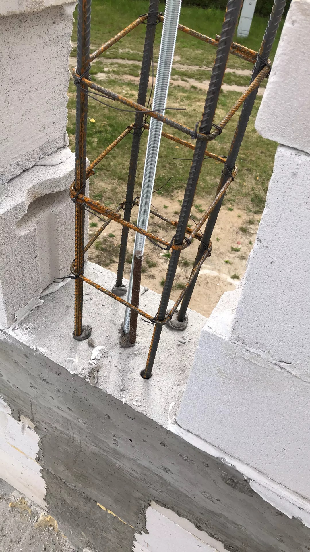 Zbrojenie pionowe z prętów stalowych, połączonych drutem wiązałkowym, wystające z fundamentu wykonanego z bloczków betonowych, z widoczną szpilką gwintowaną wewnątrz.
