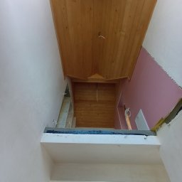 Stanisław Remonty i wykończenia - Wnętrze domu w trakcie prac wykończeniowych: drewniany sufit, ściany z płyt kartonowo-gipsowych w kolorze różowym, widoczne kable elektryczne i fragment okna. Ujęcie z dołu, ukazujące perspektywę...