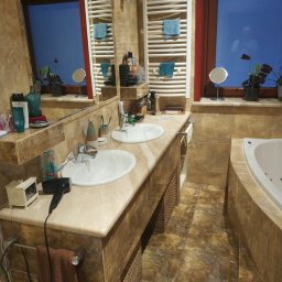 Stanisław Remonty i wykończenia - Jasna łazienka z dwoma umywalkami na kamiennym blacie, grzejnikiem drabinkowym z ręcznikami i wanną z hydromasażem, wykończona płytkami imitującymi kamień.