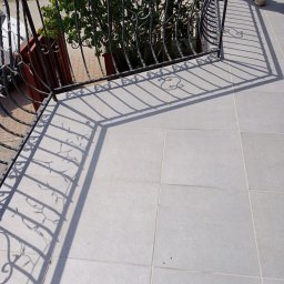 Stanisław Remonty i wykończenia - Jasnoszare płytki na balkonie z ozdobną, żeliwną balustradą rzucającą cień, widoczne buty osoby stojącej na balkonie.