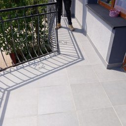 Stanisław Remonty i wykończenia - Wyłożony jasnoszarą terakotą balkon z czarną, ozdobną balustradą; widoczna osoba w ciemnych spodniach i beżowych butach.