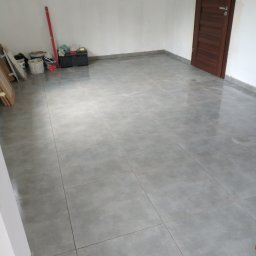 Stanisław Remonty i wykończenia - Ułożone szare płytki podłogowe w pustym pokoju z brązowymi drzwiami, widoczne narzędzia i pojemniki malarskie przy ścianie.