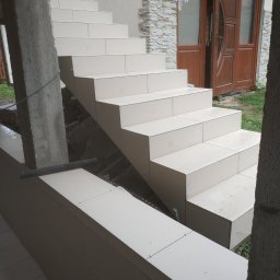 Stanisław Remonty i wykończenia - Wykończone płytkami schody zewnętrzne prowadzące do brązowych drzwi wejściowych, widoczne krzyżyki dystansowe i betonowy filar w surowym stanie.