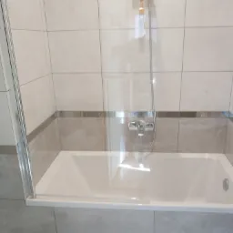 Biała wanna z chromowaną baterią i szklanym parawanem nawannowym, otoczona beżowymi płytkami w kwadraty z szarym pasem dekoracyjnym.