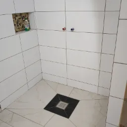 Wykończona kabina prysznicowa z białymi płytkami ściennymi, beżowymi płytkami podłogowymi z wzorem marmuru i ozdobnym odpływem liniowym. W ścianie wnęka z mozaiką i butelką płynu do mycia.