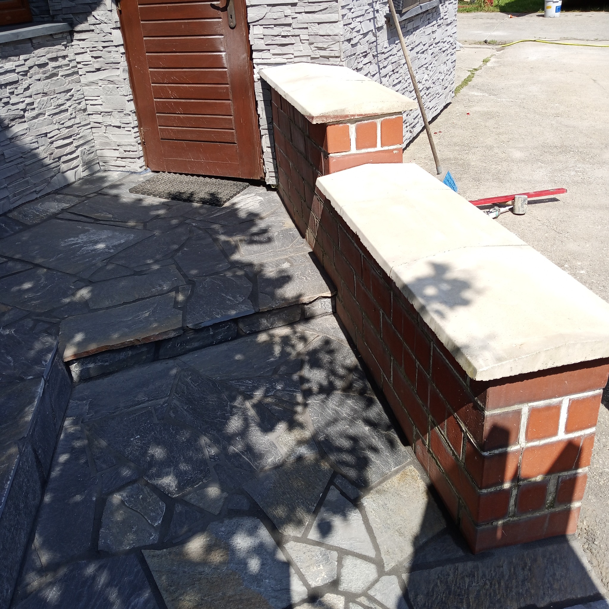 Wykończone schody wejściowe z nieregularnych, ciemnych płyt kamiennych, obok ceglane słupki z jasnymi, betonowymi nakrywami i drzwi wejściowe.