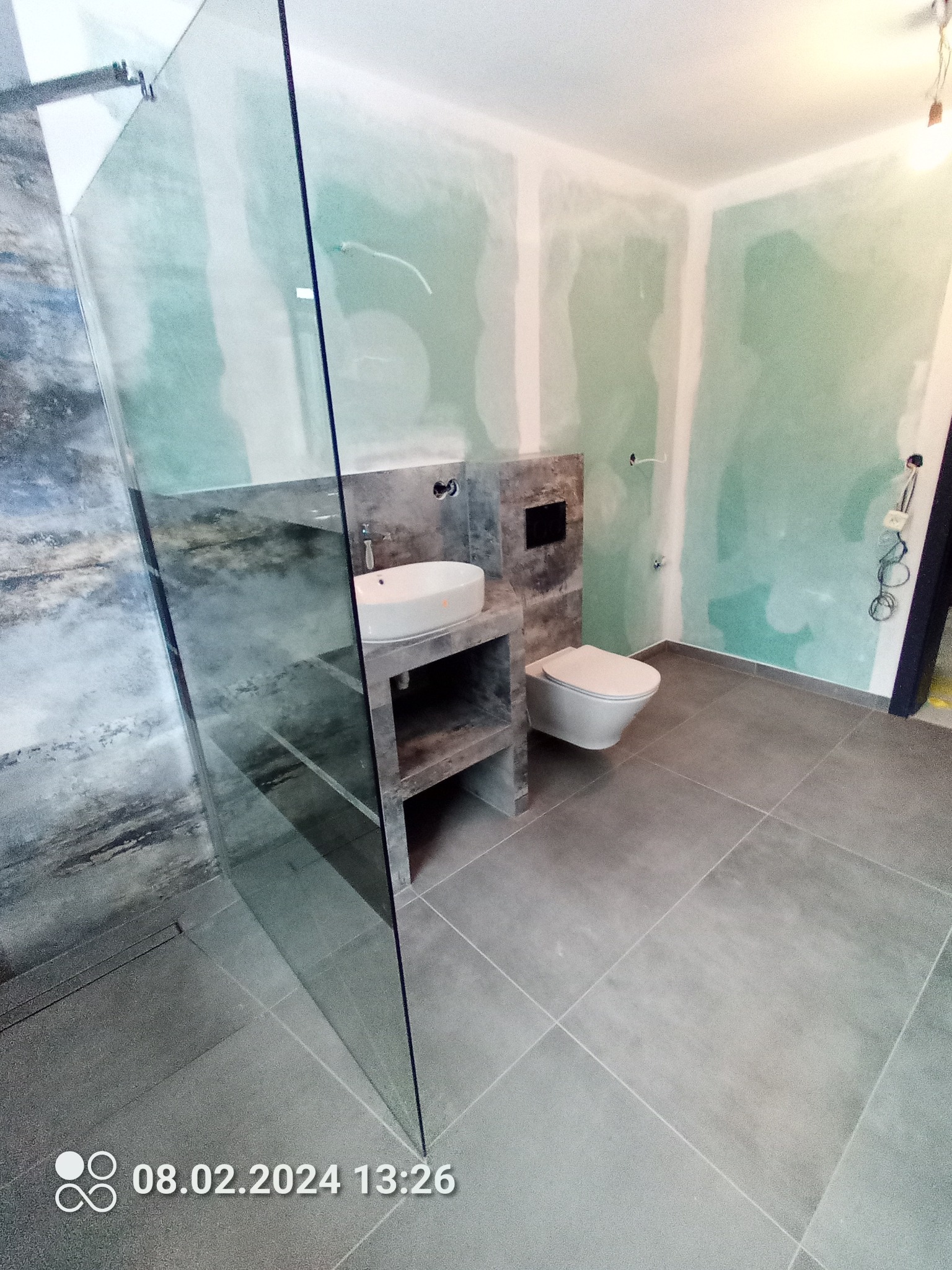 Łazienka w trakcie remontu: widoczna kabina prysznicowa ze szkła, umywalka na betonowym blacie z półkami, podwieszana toaleta, szare płytki podłogowe oraz ściany przygotowane pod gładzie.