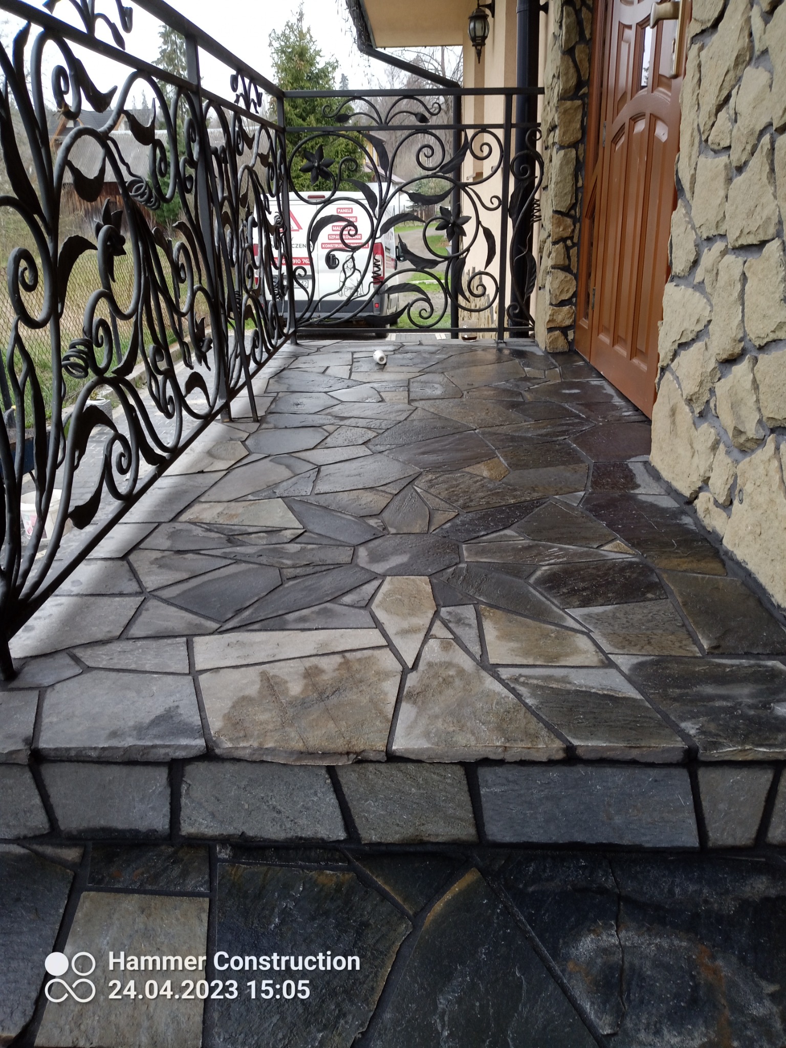 Kamienny taras z ozdobną, kutą balustradą i wejściem do domu wyłożonego kamieniem dekoracyjnym.