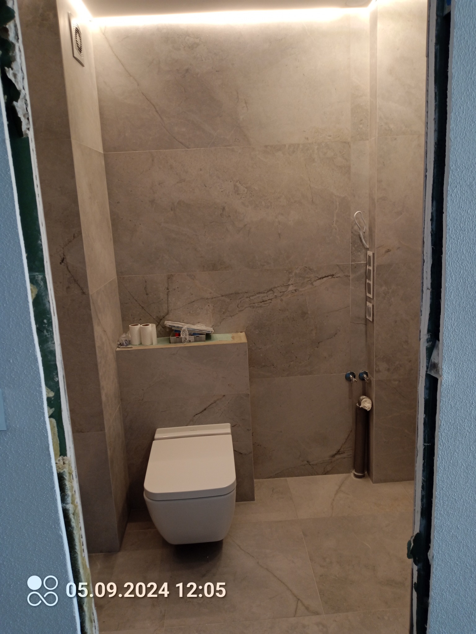 Podwieszana toaleta w trakcie montażu w łazience wyłożonej szarymi płytkami imitującymi marmur, widoczne niepodłączone rury i narzędzia na półce, oświetlenie LED podsufitowe.