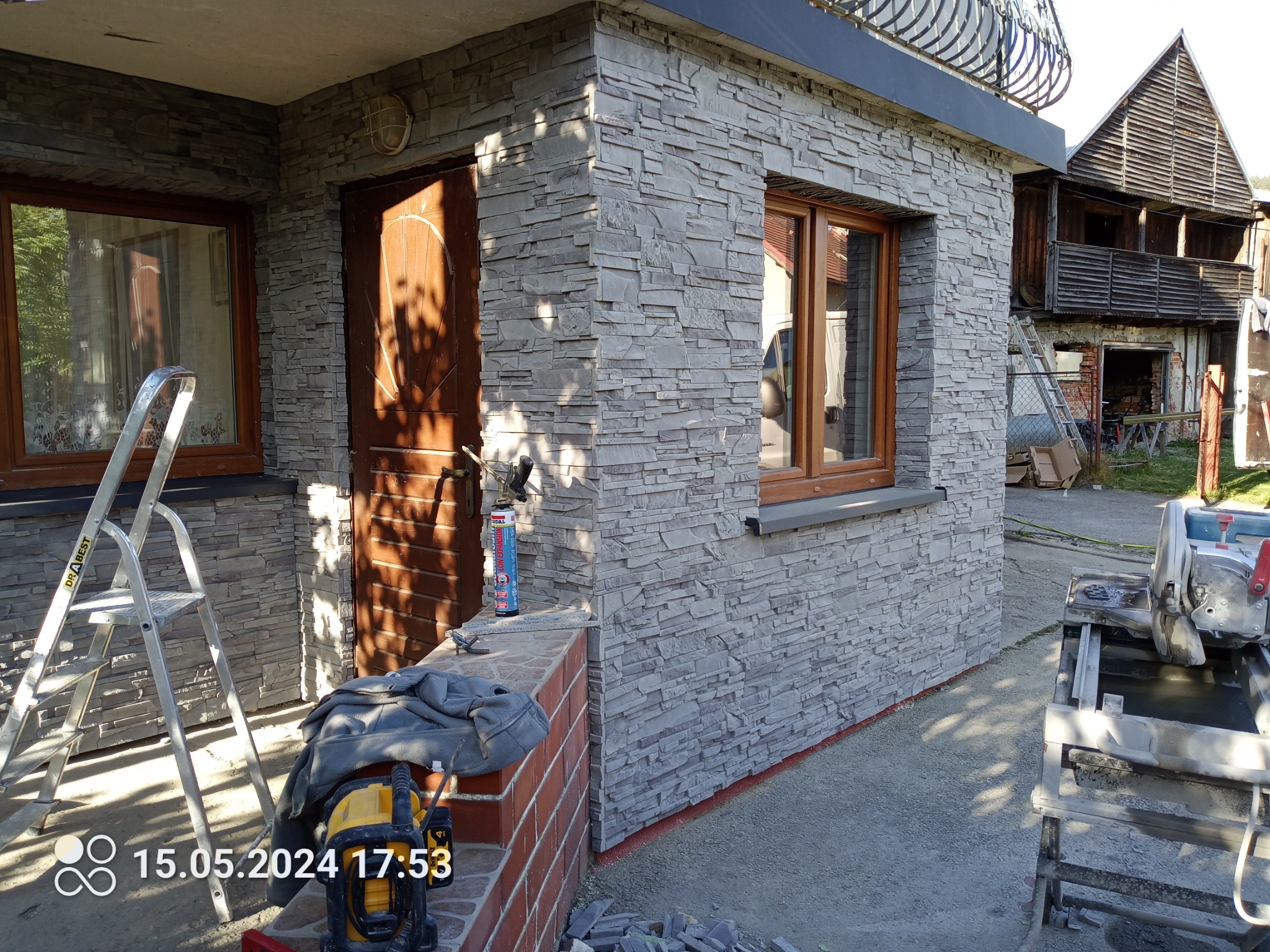 Elewacja budynku wykończona szarą okładziną imitującą kamień, widoczna drabina, narzędzia i resztki materiału budowlanego.