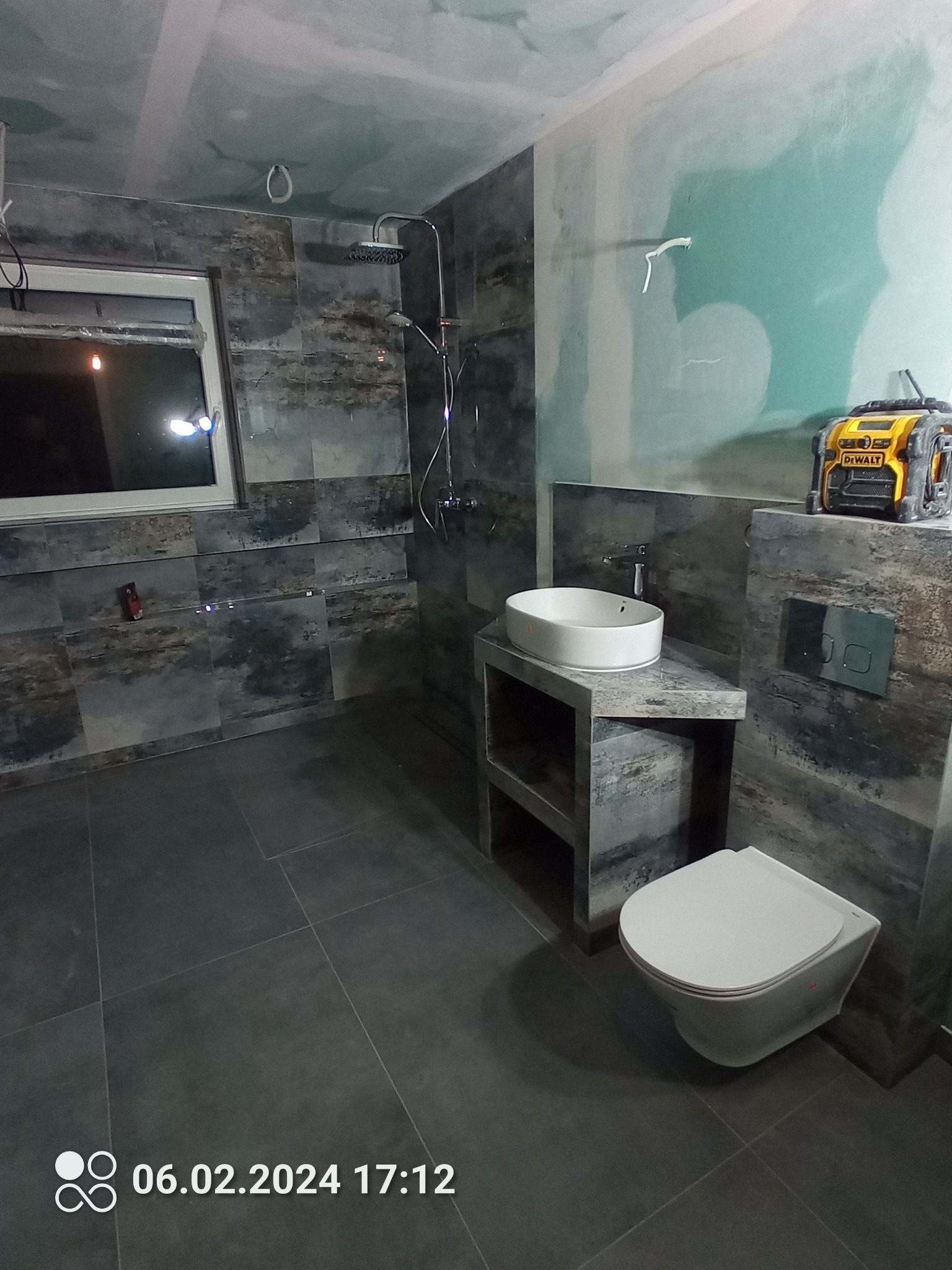 Niedokończona łazienka z szarymi płytkami imitującymi kamień, białą umywalką nablatową, toaletą podwieszaną i widoczną ścianą z płyt karton-gips, na której stoi żółty laser krzyżowy Dewalt.