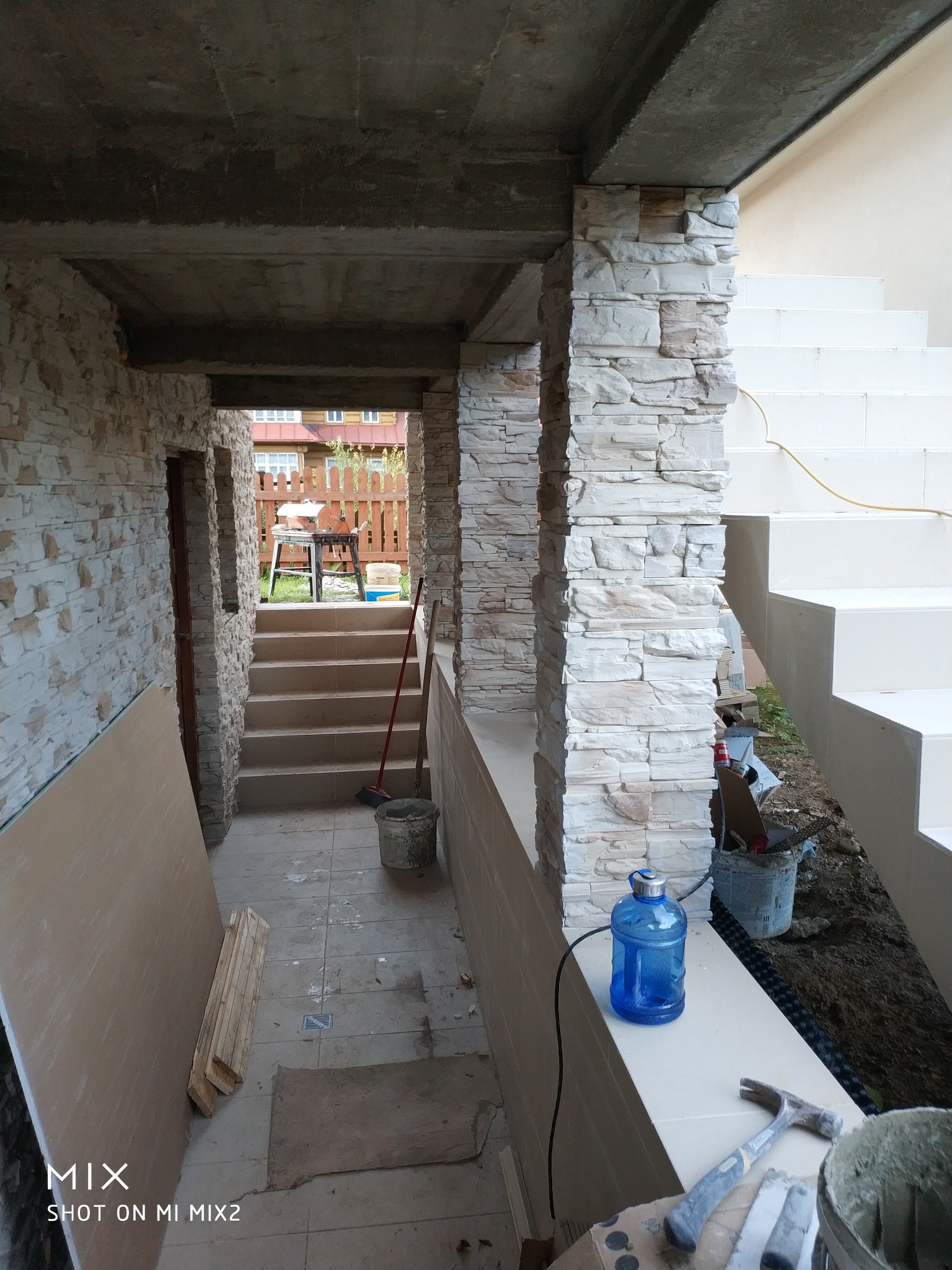 Korytarz w trakcie wykańczania z kamienną okładziną na ścianach i kolumnach, widoczne narzędzia i materiały budowlane, w tle schody i elementy konstrukcyjne budynku.