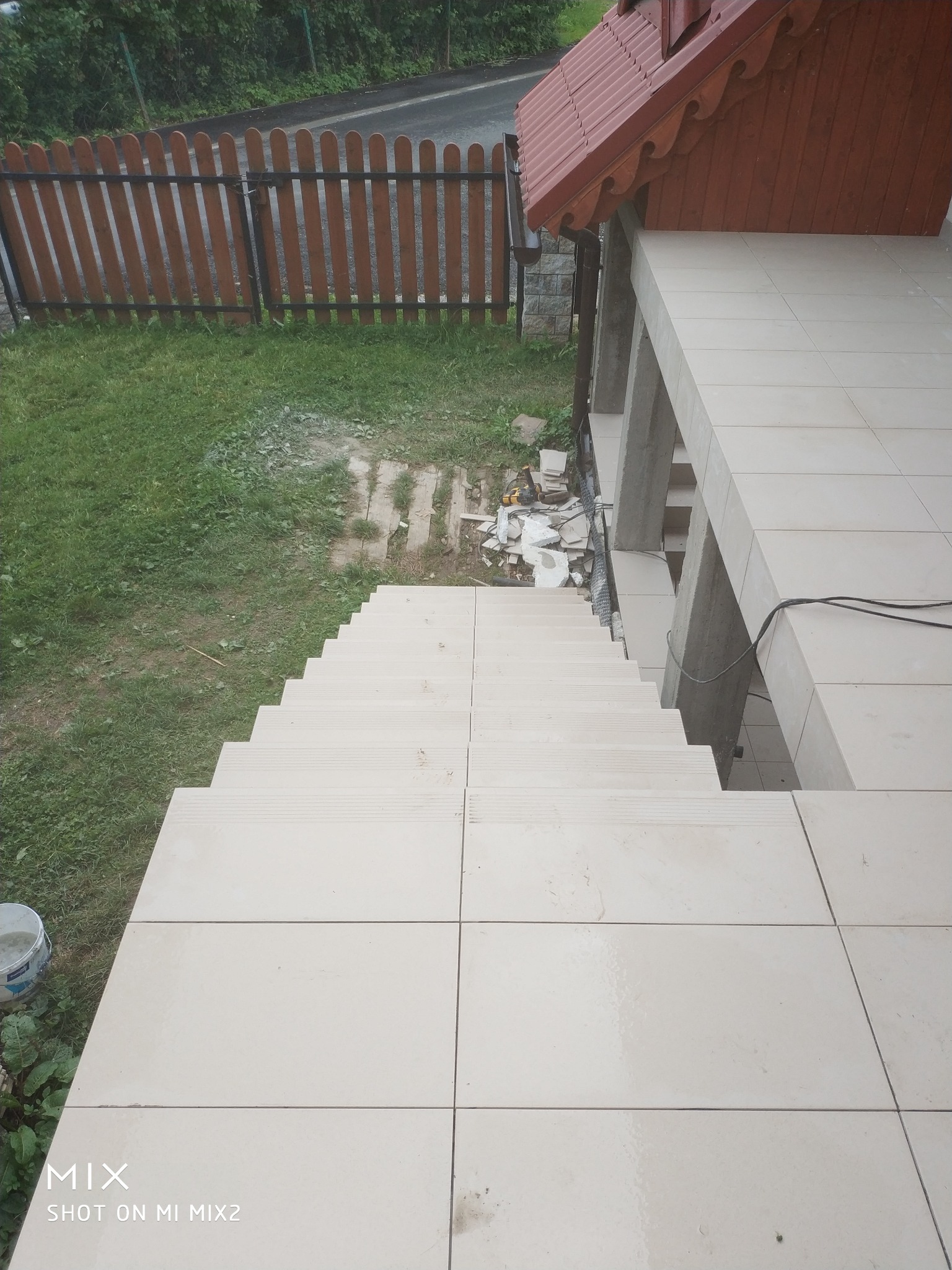 Wyłożone jasnymi płytkami schody wejściowe do domu, widok z góry, z resztkami zaprawy i narzędzi po remoncie, na zewnątrz drewniany płot i trawnik.