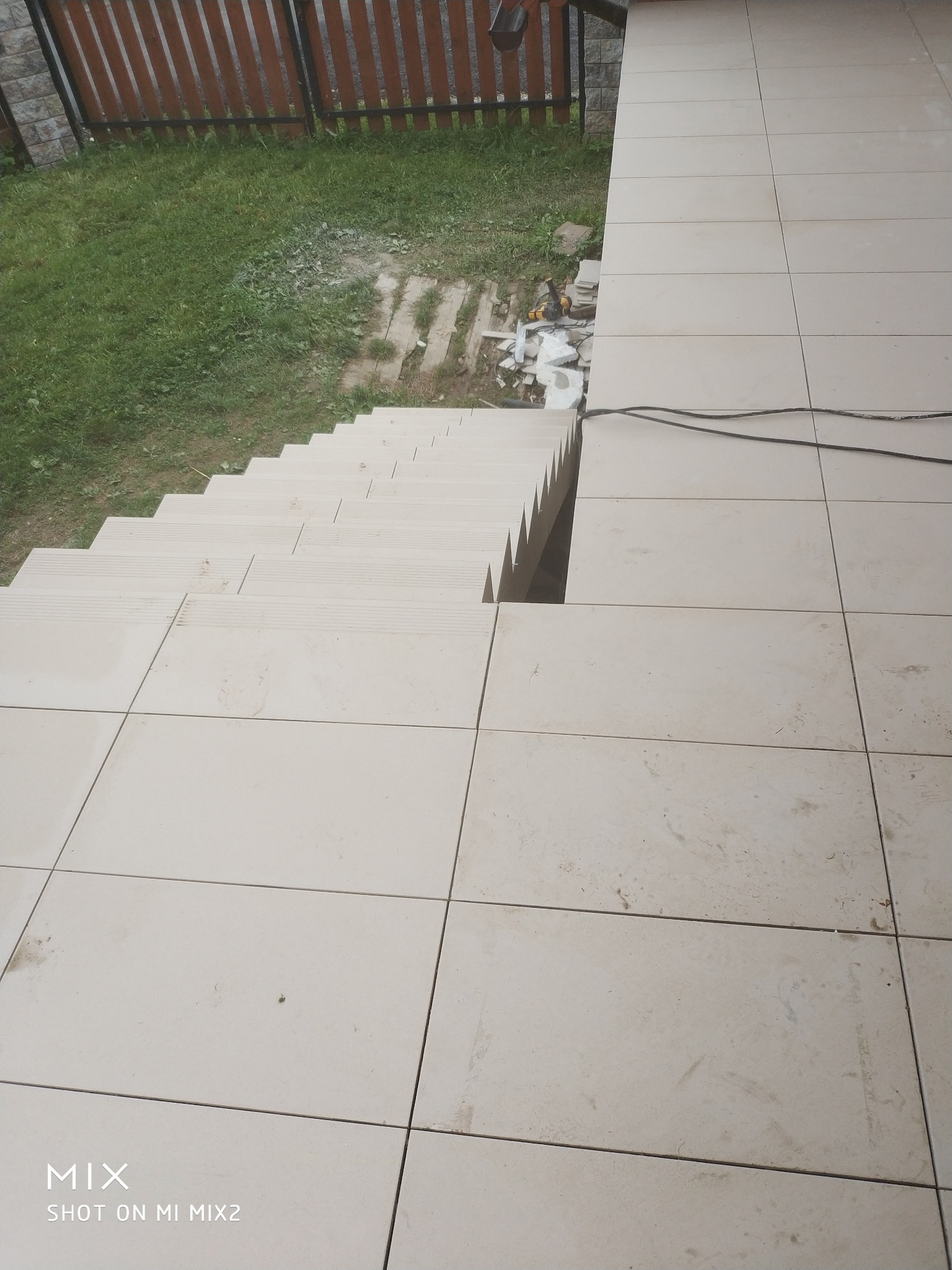 Jasne płytki ceramiczne ułożone na schodach zewnętrznych i tarasie, widoczne narzędzia glazurnicze i resztki materiałów budowlanych, brązowe ogrodzenie w tle.