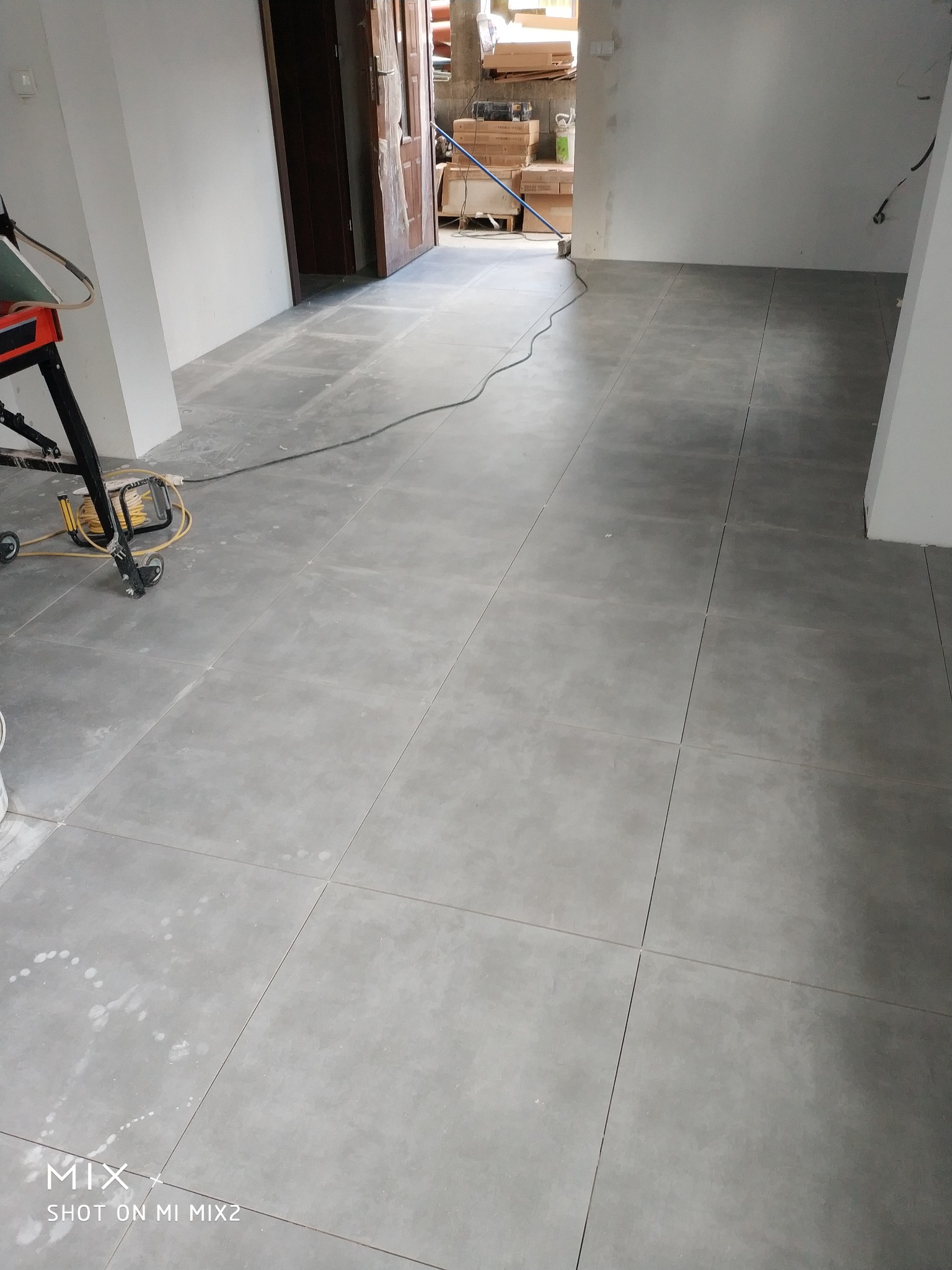 Ułożona, szara, kwadratowa podłoga z płytek ceramicznych w trakcie remontu. Widoczny sprzęt do cięcia płytek i ślady po zaprawie.