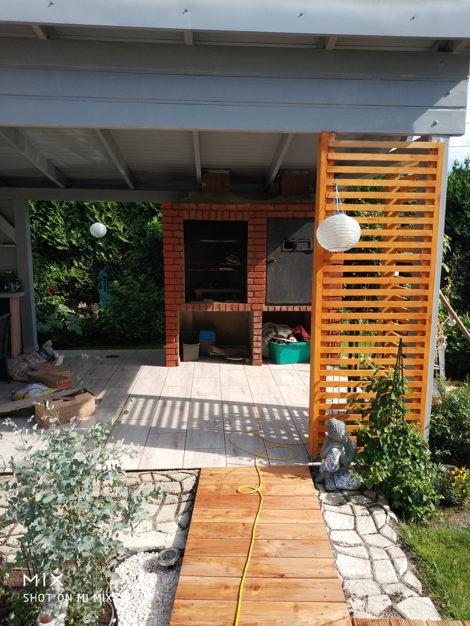 Zadaszona przestrzeń z murowanym grillem z czerwonej cegły, drewnianym ażurowym panelem, ceramiczną podłogą i drewnianą ścieżką, dekorowana lampionami i figurką Buddy.