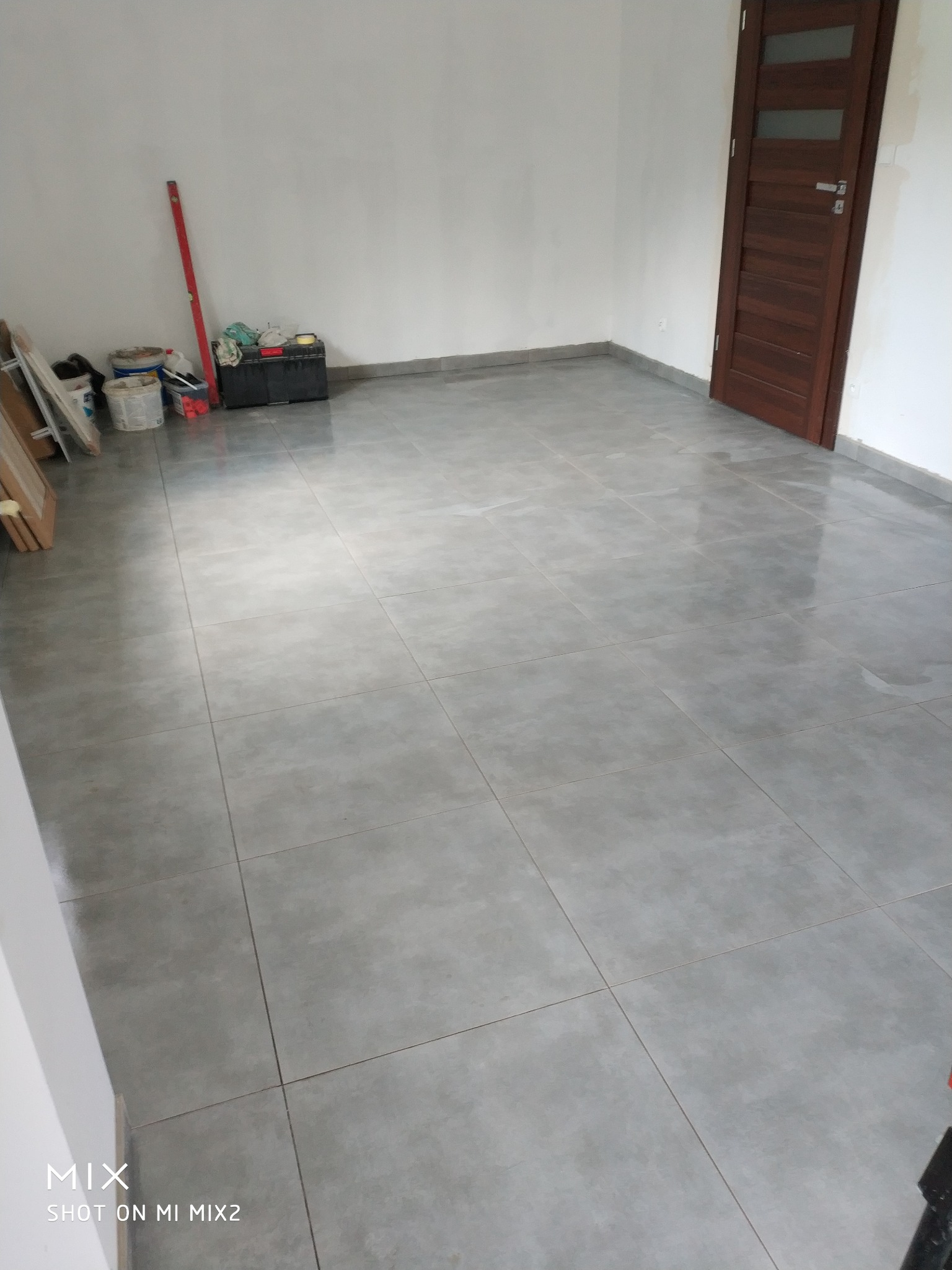 Ułożone szare płytki podłogowe w pustym pokoju z brązowymi drzwiami, widoczne narzędzia i pojemniki malarskie przy ścianie.