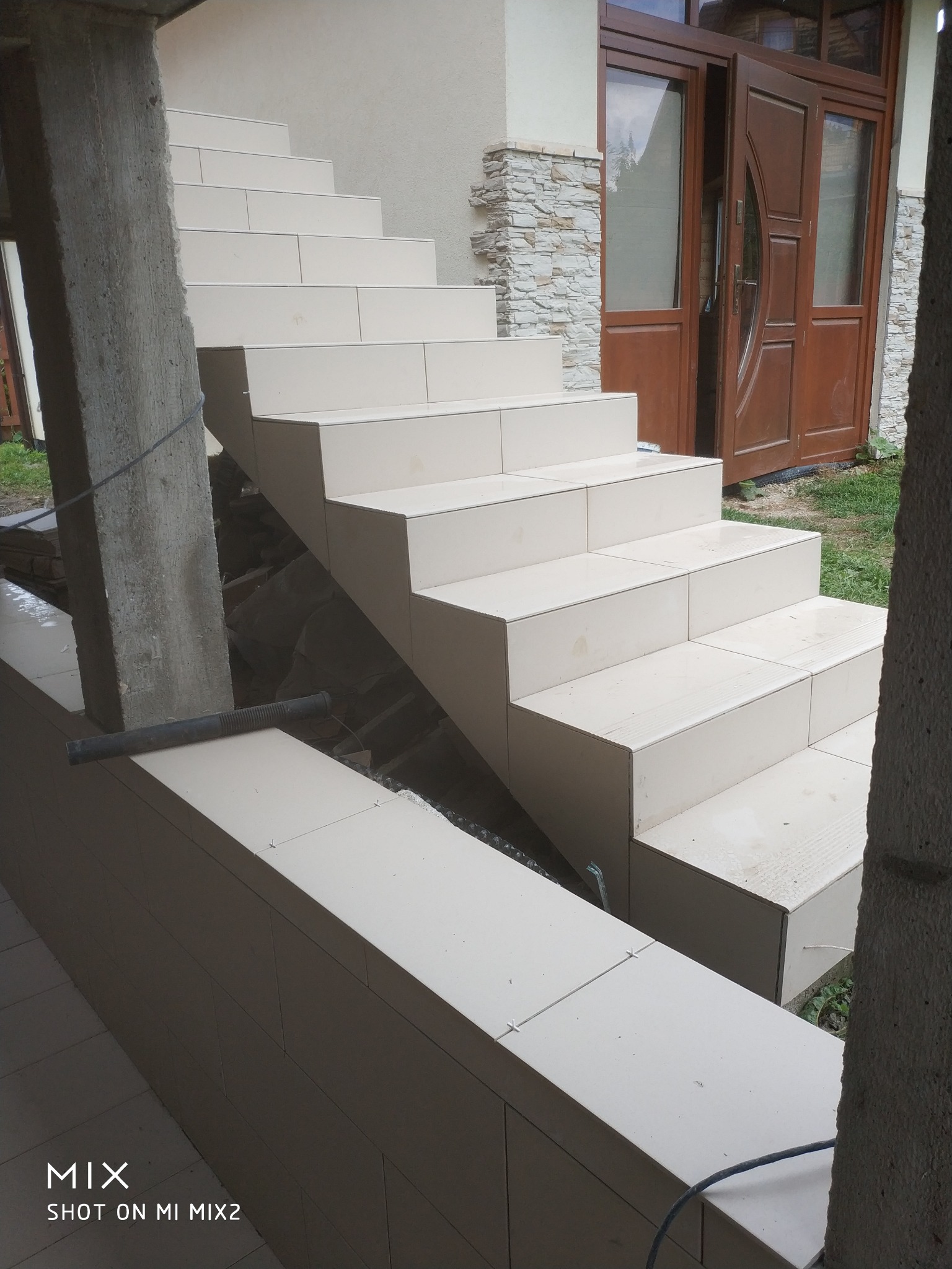 Wykończone płytkami schody zewnętrzne prowadzące do brązowych drzwi wejściowych, widoczne krzyżyki dystansowe i betonowy filar w surowym stanie.