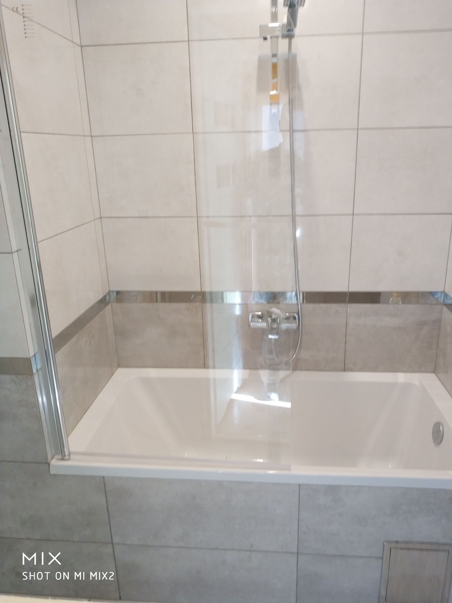 Biała wanna z chromowaną baterią i szklanym parawanem nawannowym, otoczona beżowymi płytkami w kwadraty z szarym pasem dekoracyjnym.