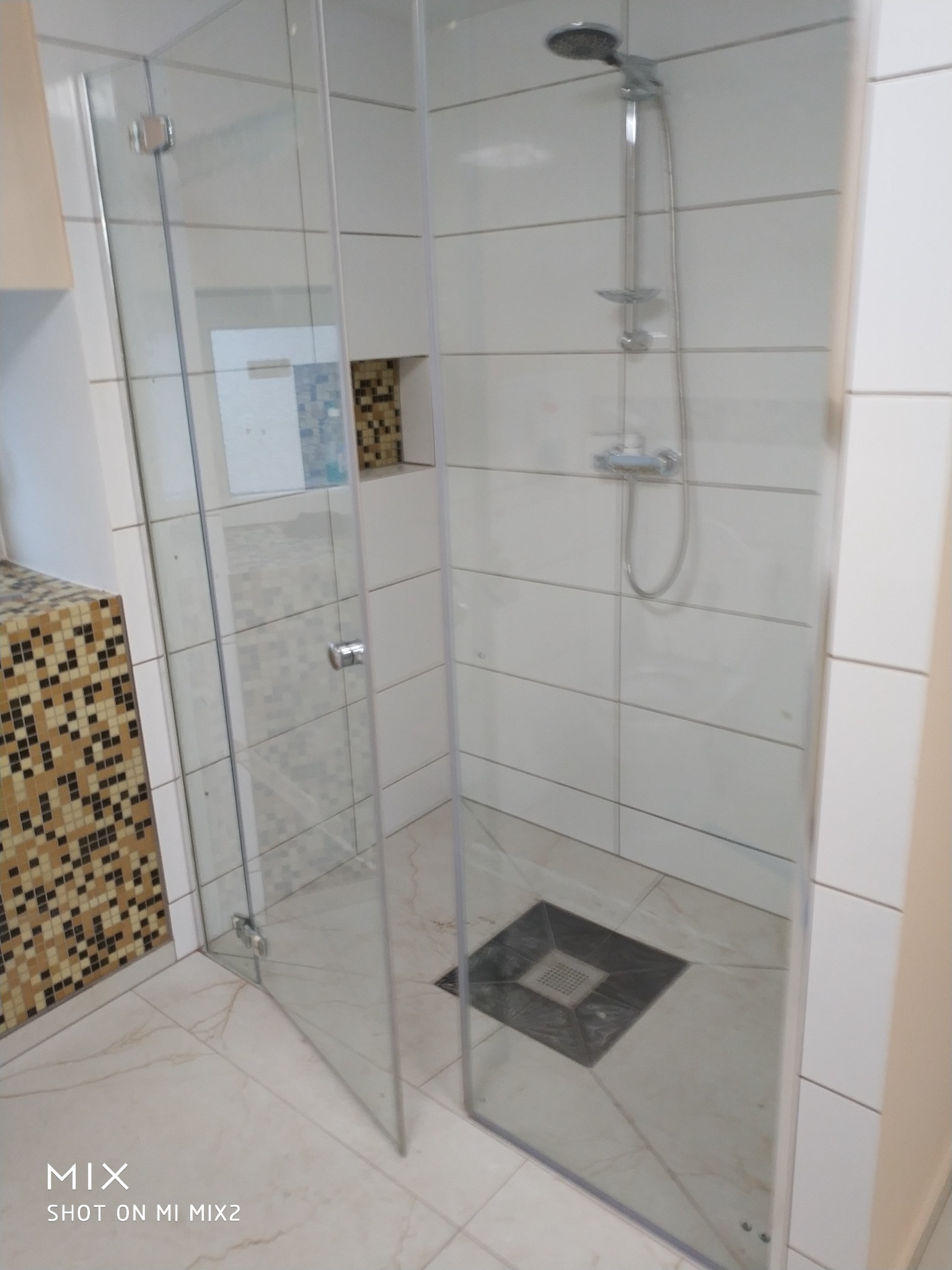 Nowoczesna kabina prysznicowa z transparentnego szkła, białe płytki ścienne z poziomymi fugami, odpływ liniowy w podłodze wyłożonej marmurkową ceramiką, fragment mozaiki dekoracyjnej obok umywalki.