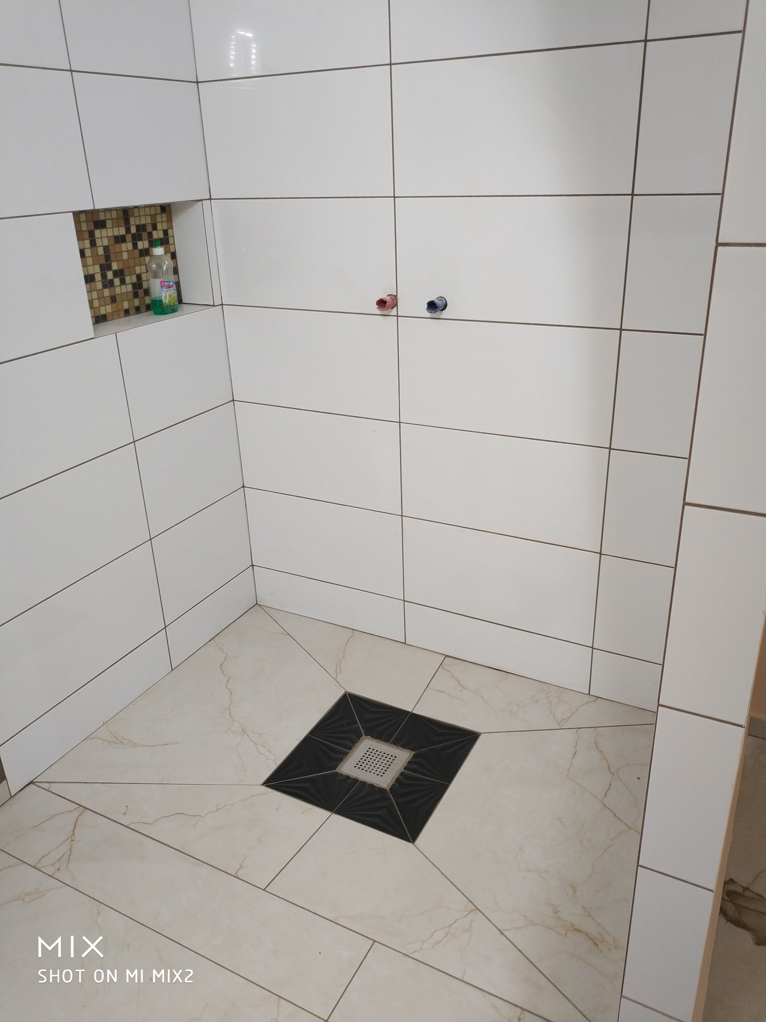 Wykończona kabina prysznicowa z białymi płytkami ściennymi, beżowymi płytkami podłogowymi z wzorem marmuru i ozdobnym odpływem liniowym. W ścianie wnęka z mozaiką i butelką płynu do mycia.